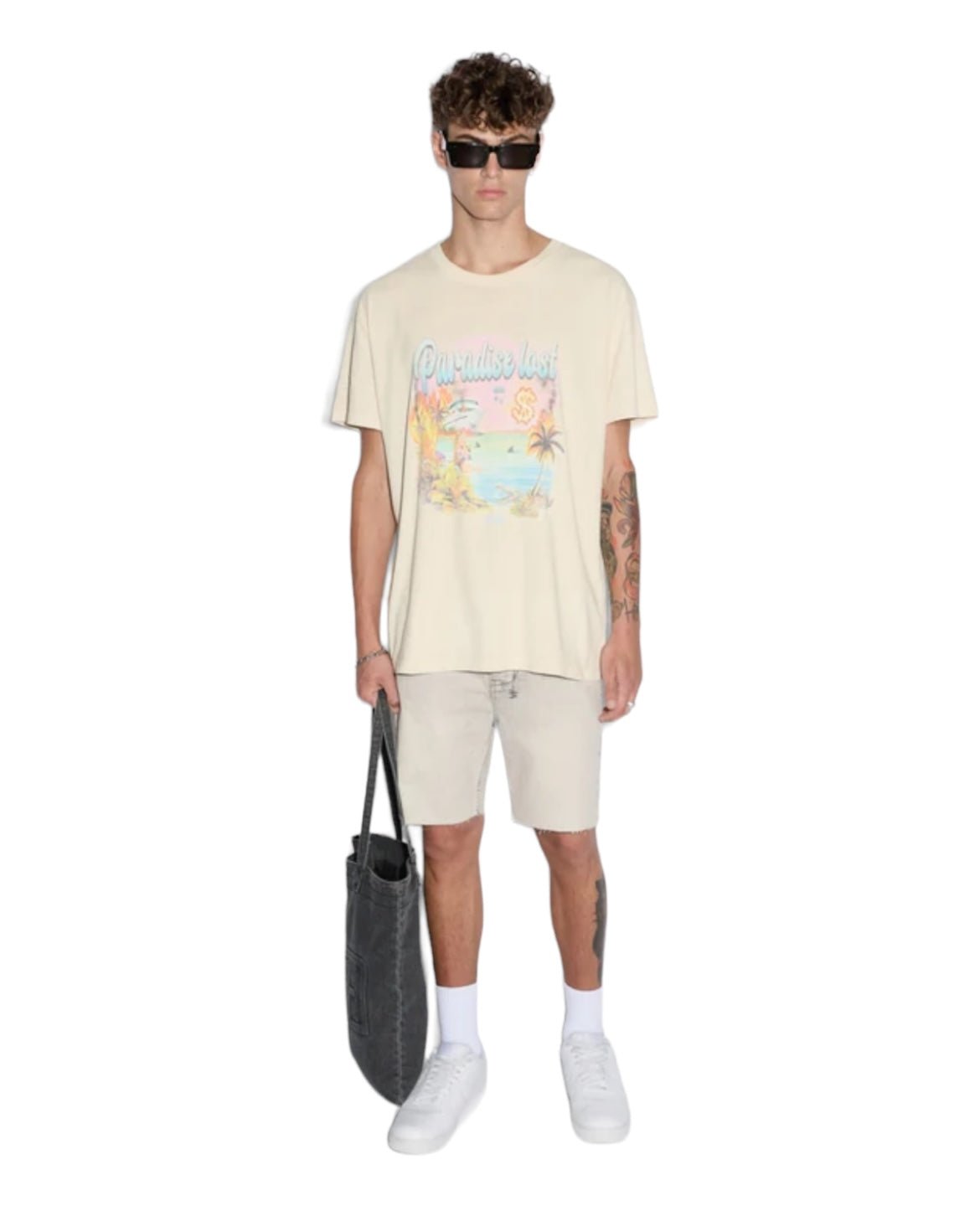 A person is wearing sunglasses, a light beige KSUBI PARADISE BIGGIE TEE DUSTY DESERT TAN with a tropical print, tan color shorts, white sneakers, and carrying a black tote bag.