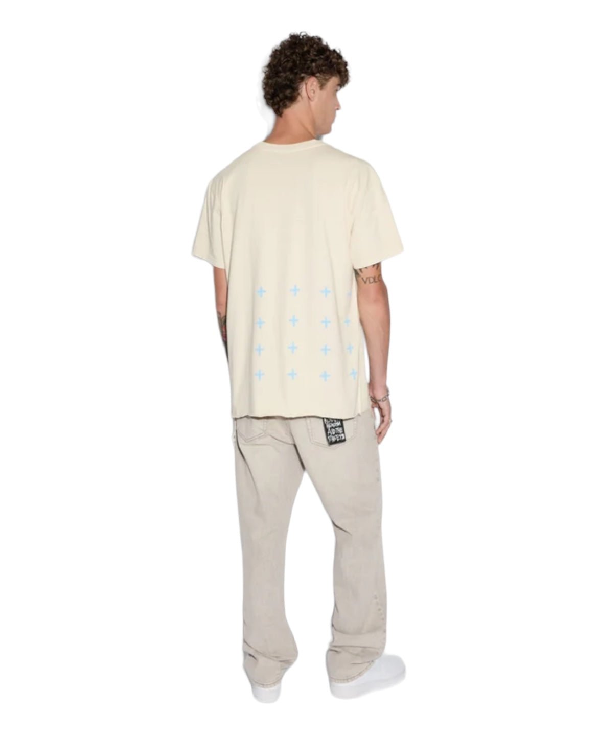 Person with curly hair wearing a KSUBI PARADISE BIGGIE TEE DUSTY DESERT TAN made of premium cotton jersey and light-colored pants, viewed from the back. Blue cross patterns are on the lower back of the t-shirt.