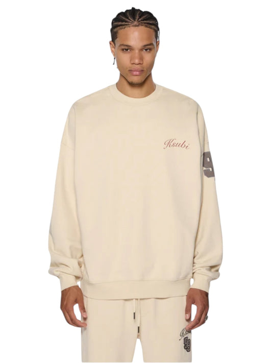 A person confidently stands against a plain background in the Ksubi Playoff Ekcess Crew Dusty Desert. This oversized, cozy crew neck sweater from KSUBI features premium fleece in a stylish beige and pairs perfectly with matching pants.