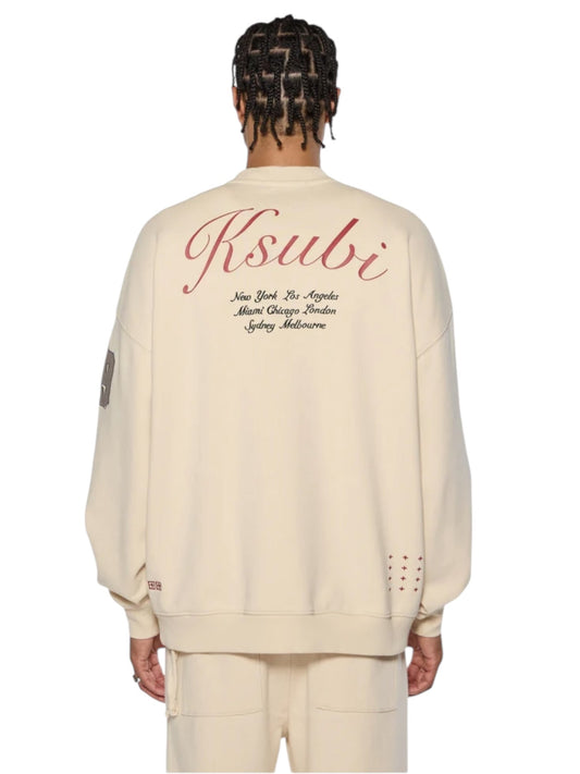 A person with braided hair wears a cream Ksubi Playoff Ekcess Crew sweatshirt in premium fleece, featuring red text on the back listing cities like New York and London, paired with matching cream pants for an effortlessly chic look.