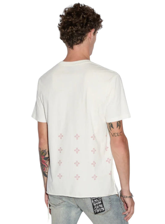 A person wearing a KSUBI SINNERS KASH SS TEE in vintage white, paired with light blue jeans. The shirt features red plus signs near the bottom on the back. The person has tattoos on both arms and holds a keychain from their back pocket, subtly showcasing their unique style.