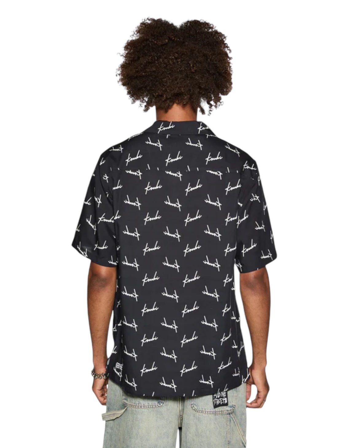 A person with curly hair is seen from the back wearing a short-sleeve, black KSUBI SKRIPT RESORT SS SHIRT featuring white text and design patterns, paired with light gray pants.