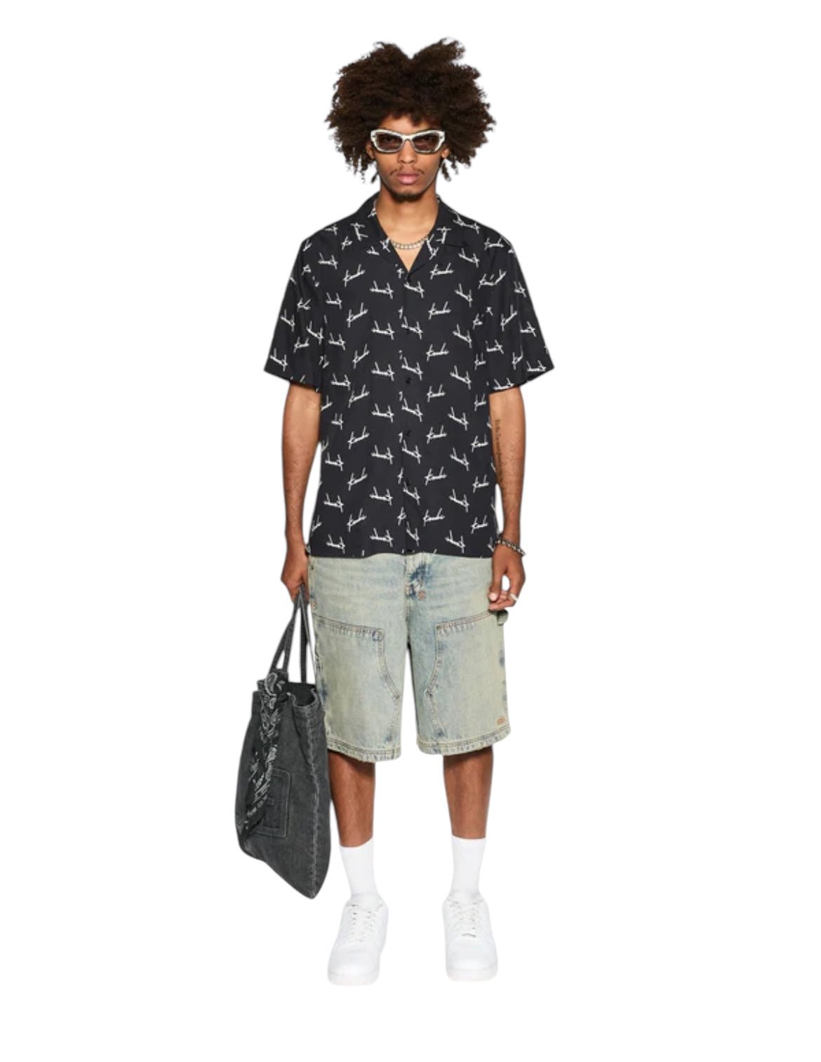 A person is standing, wearing sunglasses, a KSUBI SKRIPT RESORT RESORT SS SHIRT in black with white patterns, denim shorts, and white sneakers. They are holding a black bag featuring subtle Ksubi branding.