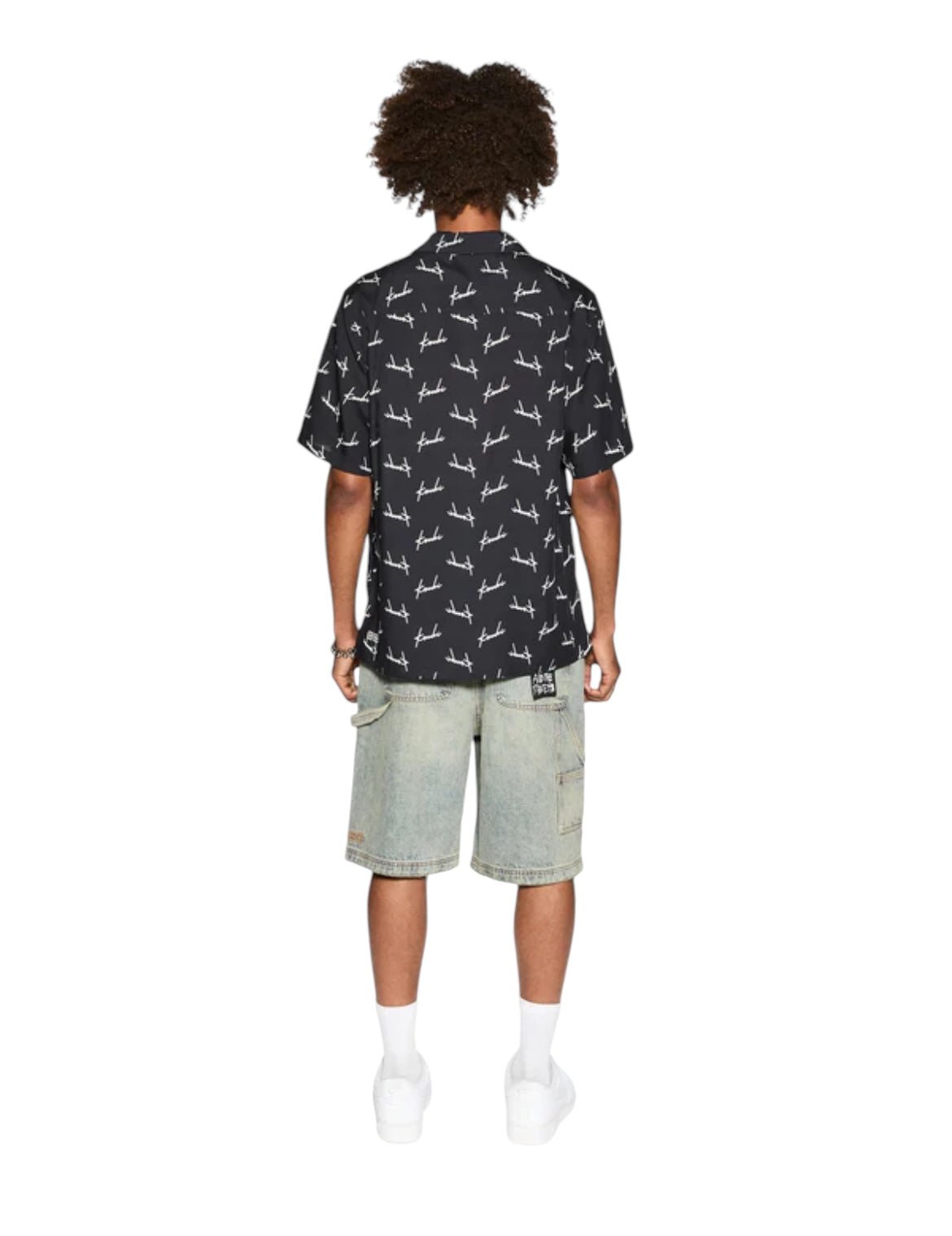 A person with curly hair stands facing away, wearing a short-sleeve KSUBI Skript Resort SS Shirt in black featuring white graphics, light blue denim shorts, white socks, and white shoes.