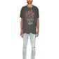 A person stands facing forward in a Ksubi Skull Biggie Ss Tee Faded Blk from KSUBI, showcasing a flaming skull graphic. They pair it with light ripped jeans and white shoes, all set against a plain white backdrop.