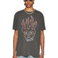 A person in a Ksubi Skull Biggie Ss Tee Faded Blk by KSUBI, oversized with a flaming skull design and premium cotton feel, accessorized with sunglasses, a chain necklace, and blue jeans stands confidently against a plain white background.