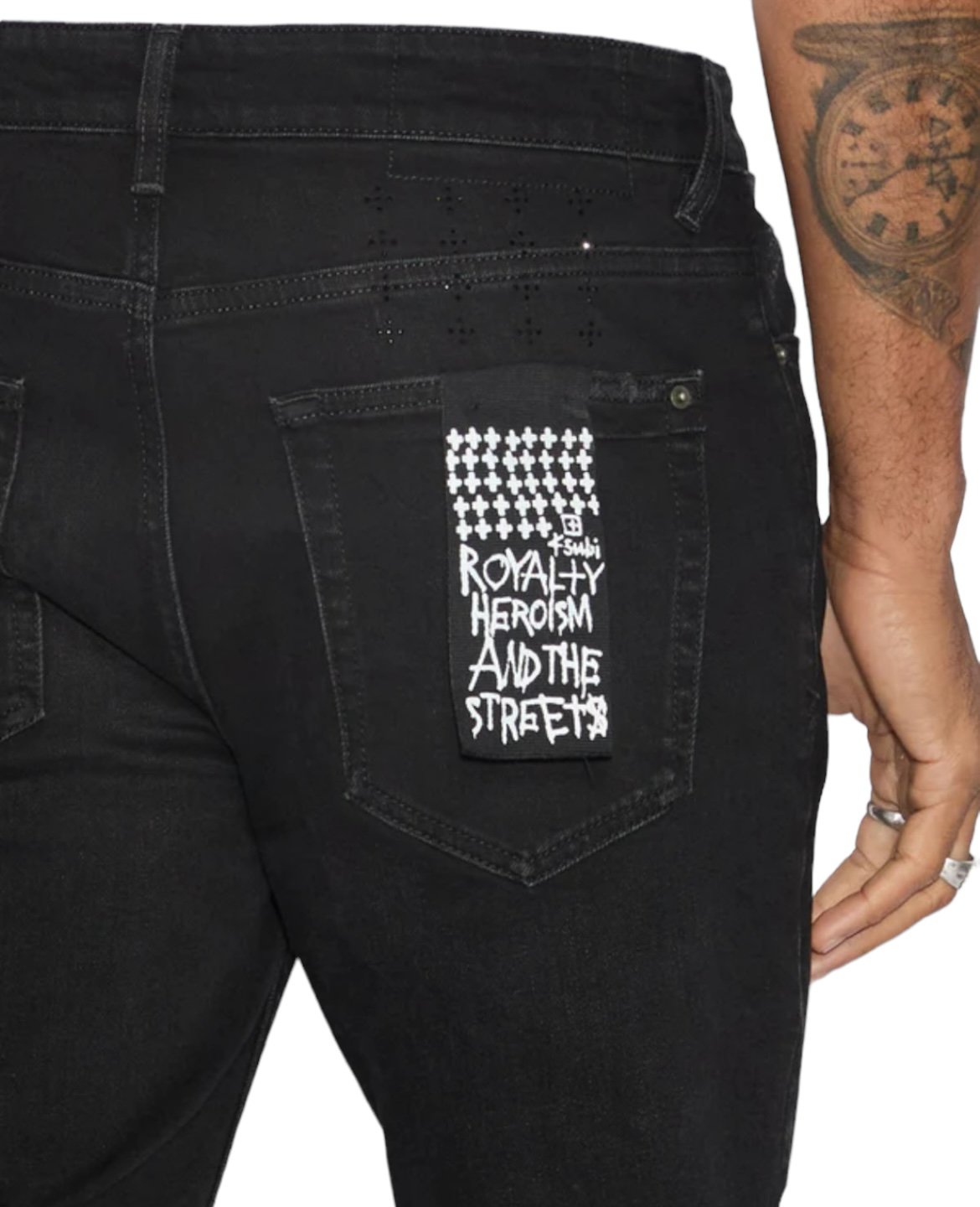 A person wearing KSUBI VAN WINKLE APEX KRYSTAL REPAIR BLACK jeans with a black and white patch on the back pocket that reads "Royalty Heroism and the Streets." Their left arm, with a clock tattoo, and hand with a ring are visible.