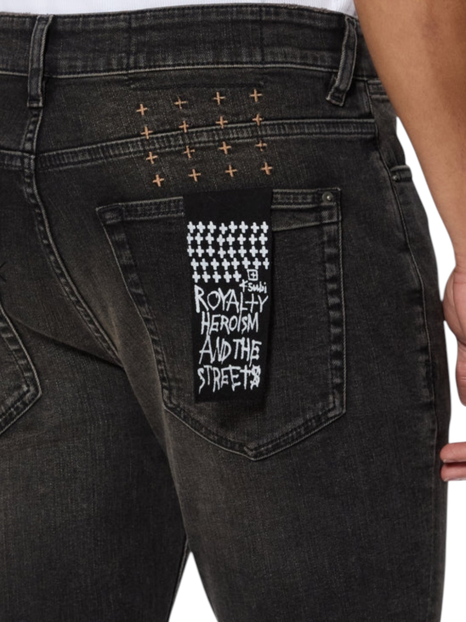 Close-up of the back pocket on KSUBI Van Winkle Kingdom Repair Black skinny jeans, showing a patch with "Royalty Heroism And The Streets" and a pattern of white Ksubi cross logos above the pocket.