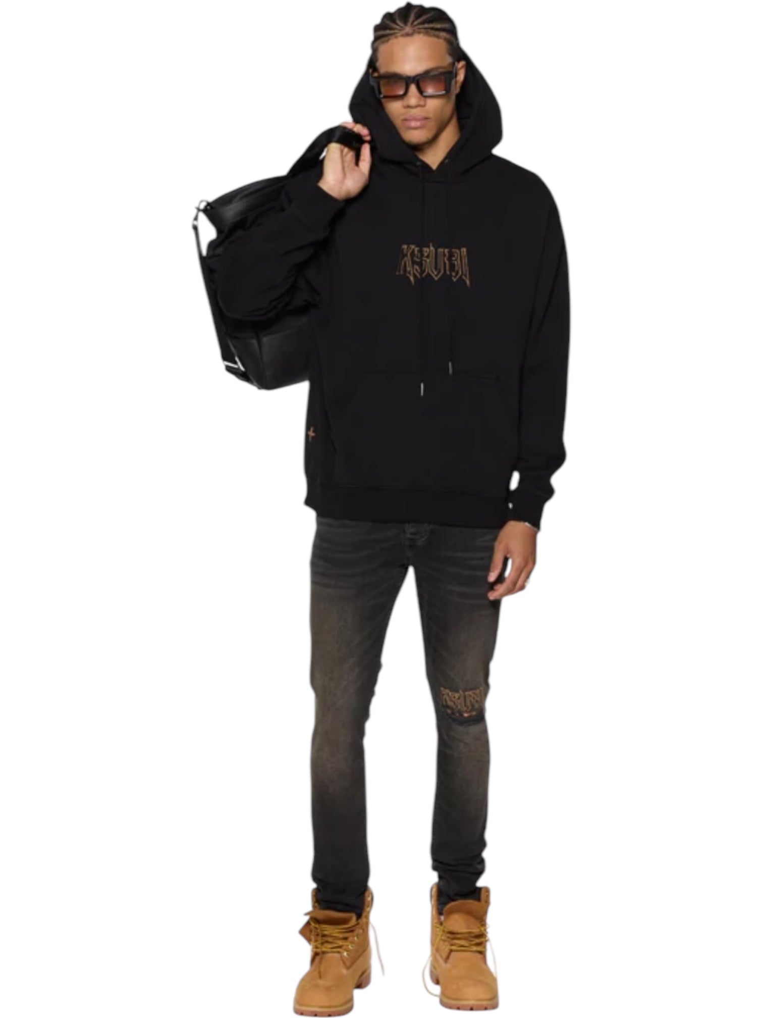 A person stands against a white background, wearing a black hoodie, tan boots, sunglasses, carrying a large black bag, and KSUBI's Van Winkle Kingdom Repair Black denim skinny jeans.