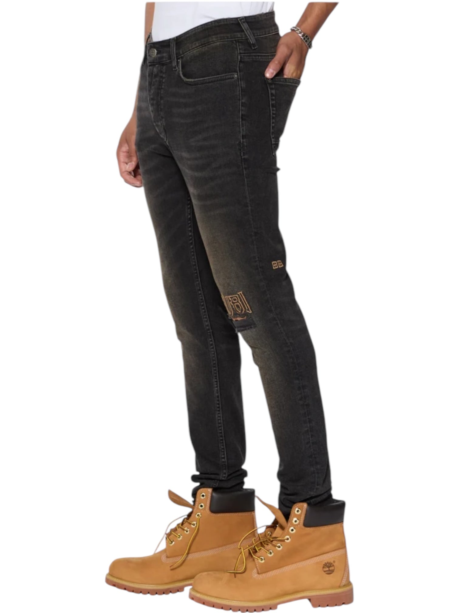 A person in KSUBI Van Winkle Kingdom Repair Black skinny jeans with a knee patch and tan lace-up work boots with the Ksubi logo stands against a white background.