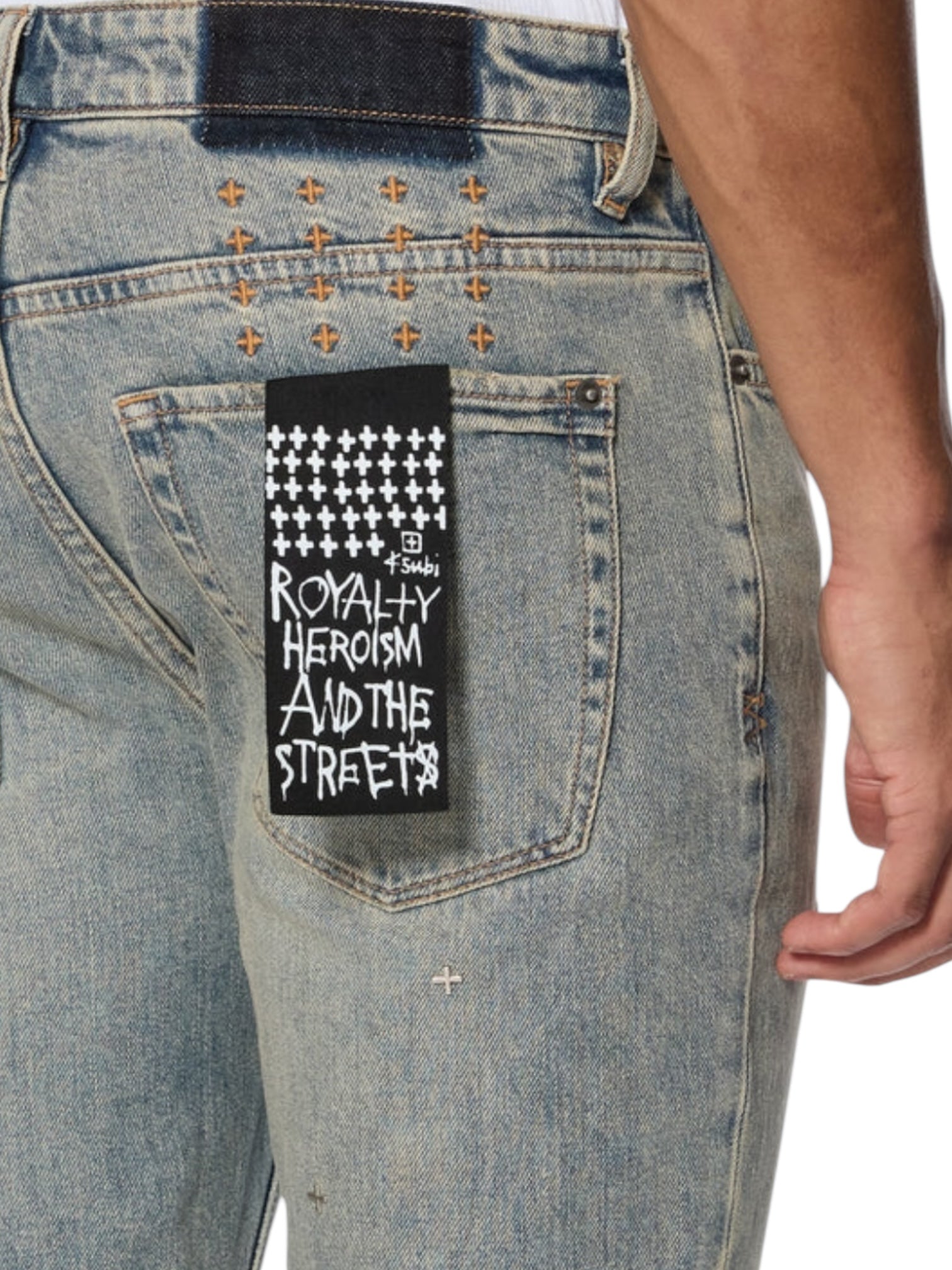 Wearing KSUBI Van Winkle Stagelight Metalik Denim—premium light blue skinny jeans with gold cross-stitching, a black back pocket patch reading "ROYALTY HEROISM AND THE STREETS," and Ksubi-branded hardware.