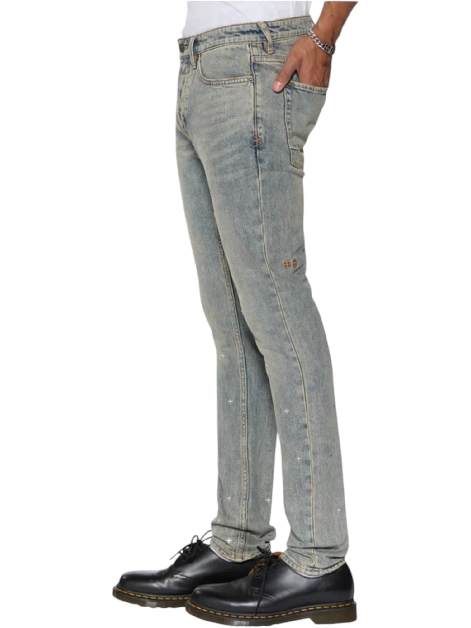 A person in KSUBI Van Winkle Stagelight Metalik Denim jeans, a white shirt, and black lace-up shoes stands in side profile against a white background.