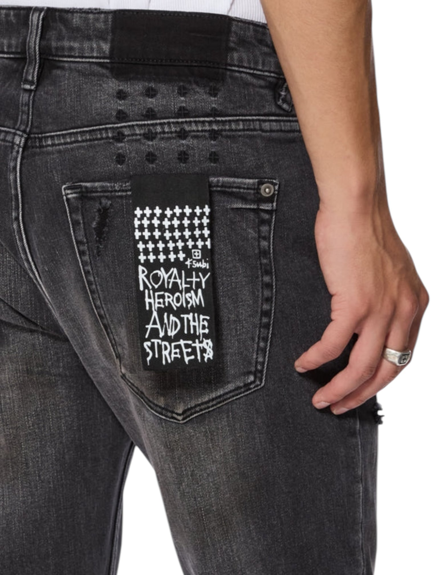 Close-up of the back pocket on KSUBI's ksubi Van Winkle Torqued Chamber Black jeans, featuring a "ROYALTY HEROISM AND THE STREETS" patch and worn by someone with a ring, highlighting premium comfort stretch.