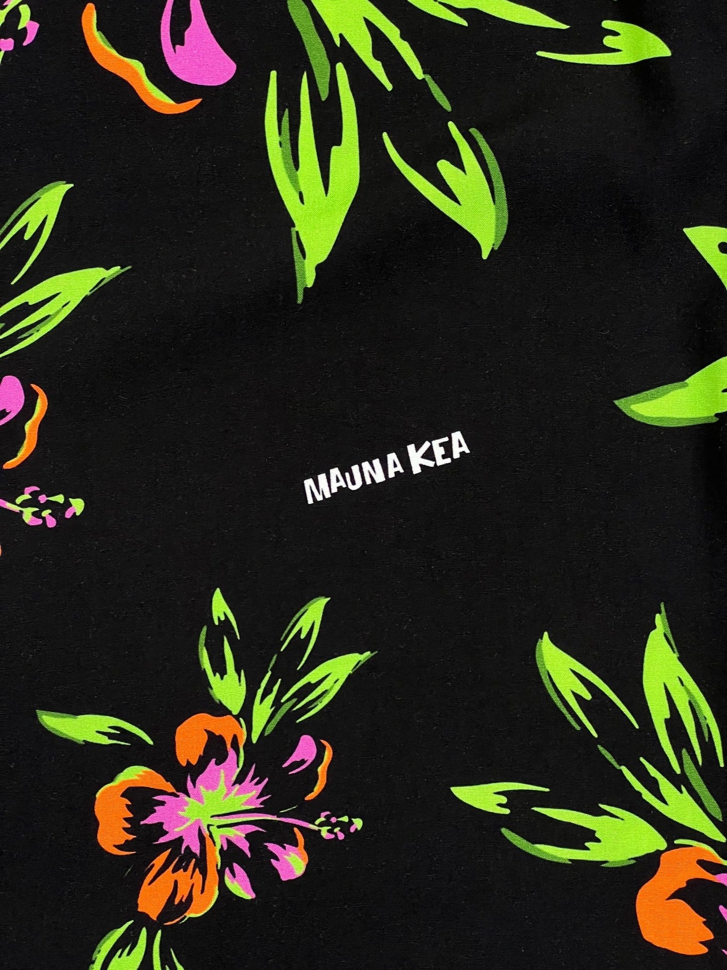 A close-up of a MAUNA-KEA MKU142-FL floral shirt in vibrant green, orange, and pink colors on a black background. The text "mauna kea" is printed in white.