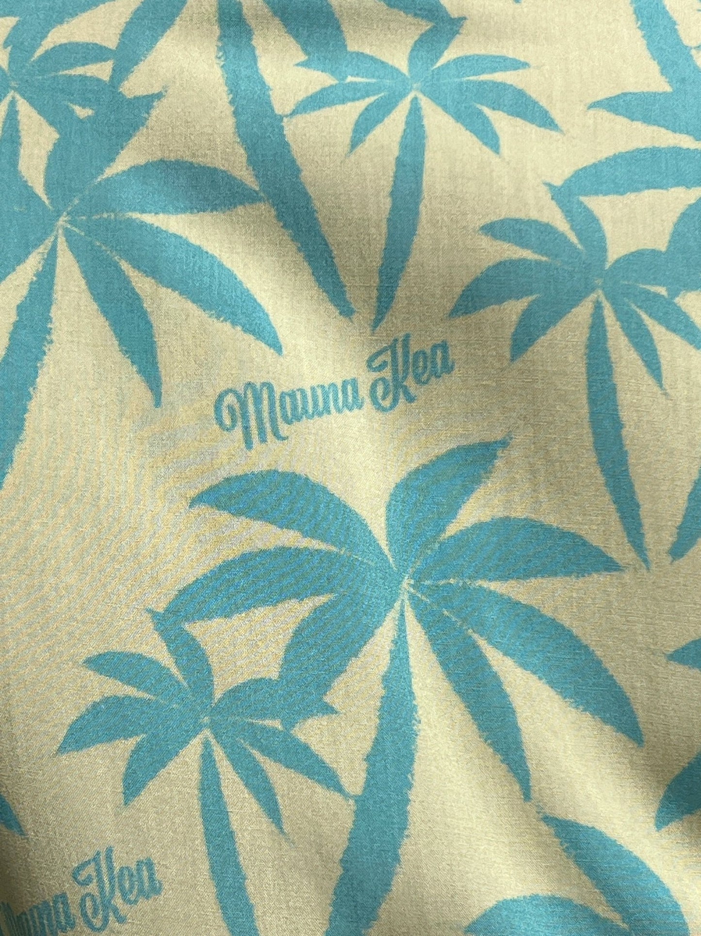 A short sleeve MAUNA-KEA MKU142-PM2 ALLOVER PRINT SHIRT GRN featuring a Mauna-Kea graphic with blue palm tree prints and the text "Mauna Kea" on a light beige background, proudly Made in Italy by MAUNA-KEA.