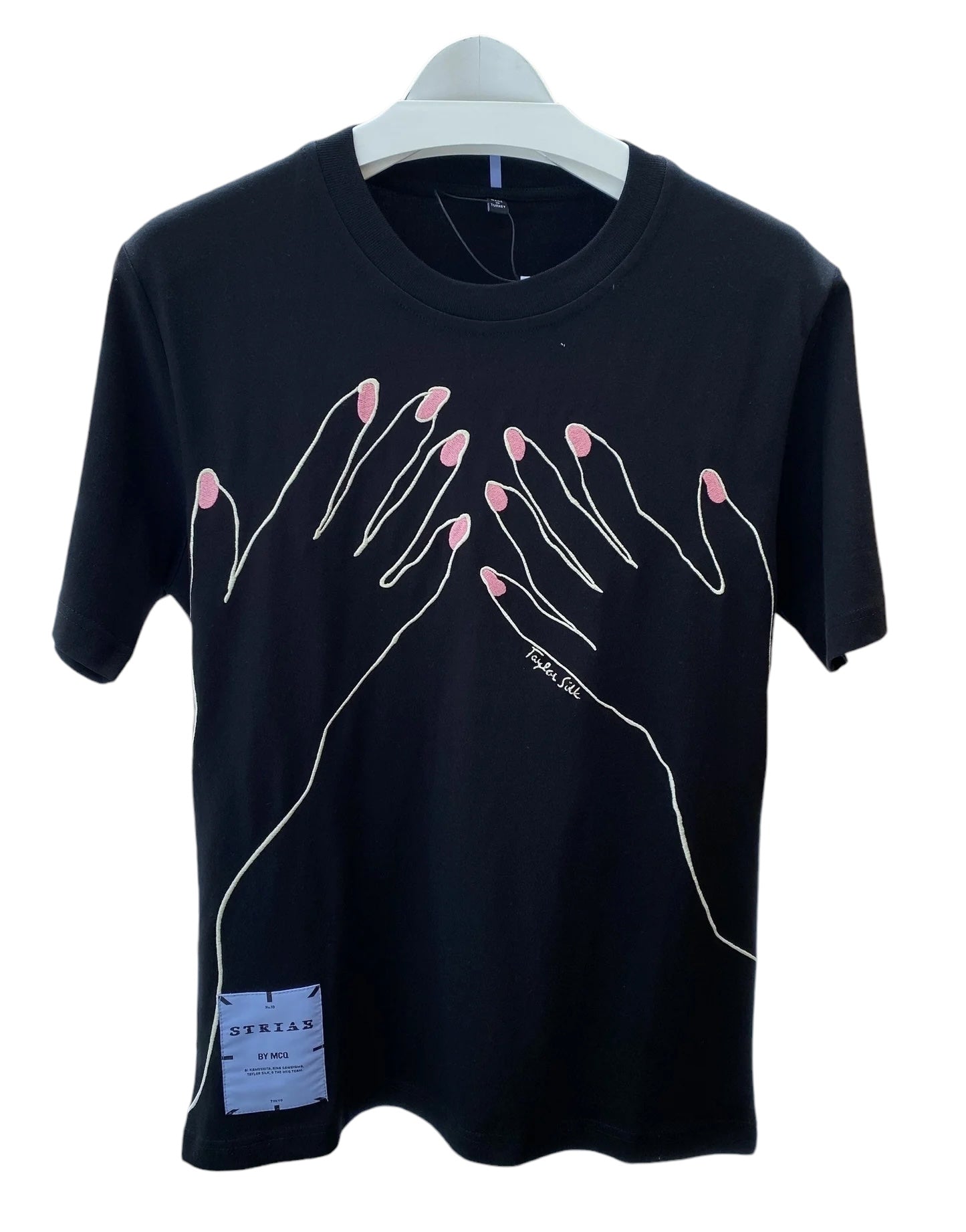 Display of a Mcq By Mcqueen 647244Rst87 T-Shirt in darkest black featuring a two-hand illustration with pink nails. A small tag subtly adorns the embroidered graphic design, enhancing the visual appeal on the hanger. Brand: MCQ BY MCQUEEN.