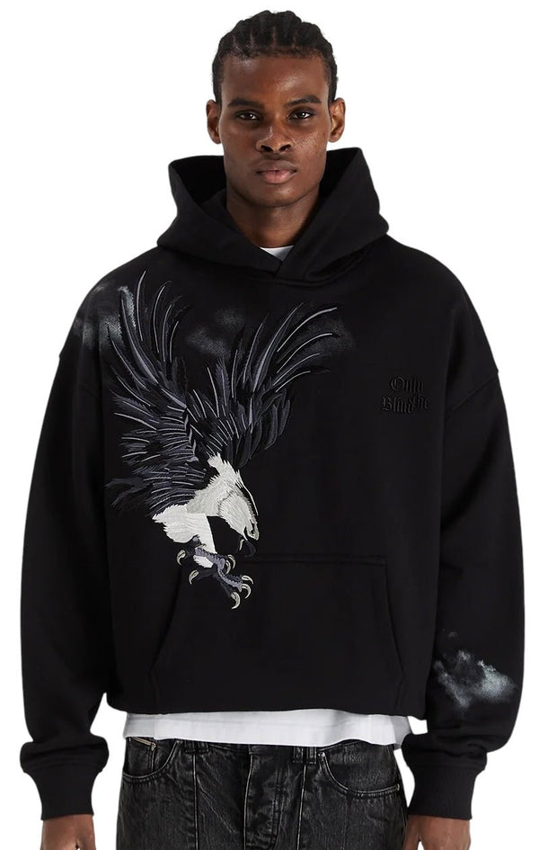 Sporting the Only The Blind OTB-H1355 Midnight Hawk Hoodie Blk, featuring an eagle graphic and crafted from GOTS® Certified Organic Cotton, this individual pairs it with dark pants. The hoodie’s detailed embroidery highlights style and eco-consciousness.