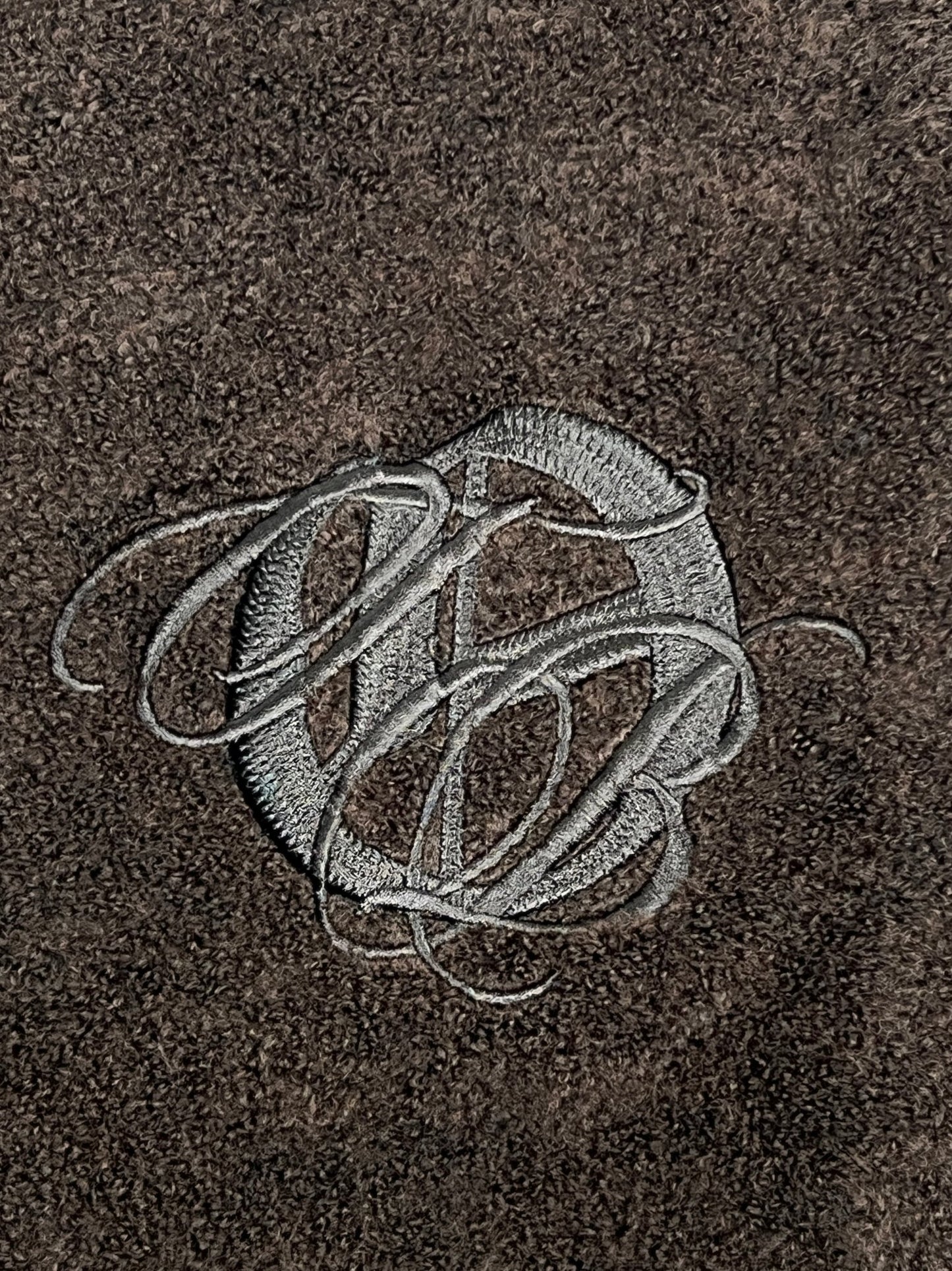 Gray embroidered script letters form a monogram on textured dark brown fabric, inspired by the ONLY THE BLIND OTB-K1629 Brown Studio Dept. Cardigan.