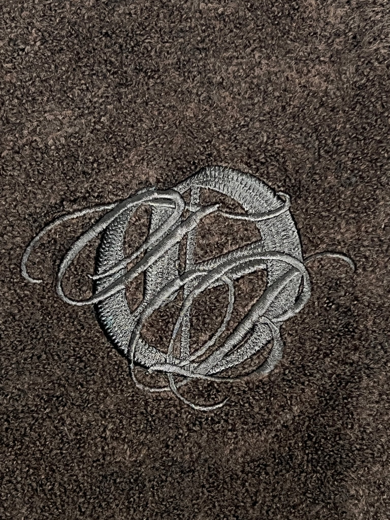 Gray embroidered script letters form a monogram on textured dark brown fabric, inspired by the ONLY THE BLIND OTB-K1629 Brown Studio Dept. Cardigan.