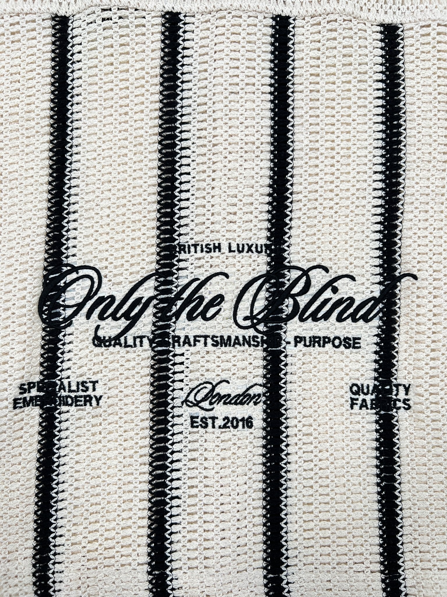 The Desert Stripe Crochet Shirt by ONLY THE BLIND features beige-black vertical stripes with text: "Only the Blind, British Luxury, Quality Craftsmanship & Purpose, London, Est. 2016." Renowned for expert embroidery and premium fabrics, it epitomizes sophistication and style.