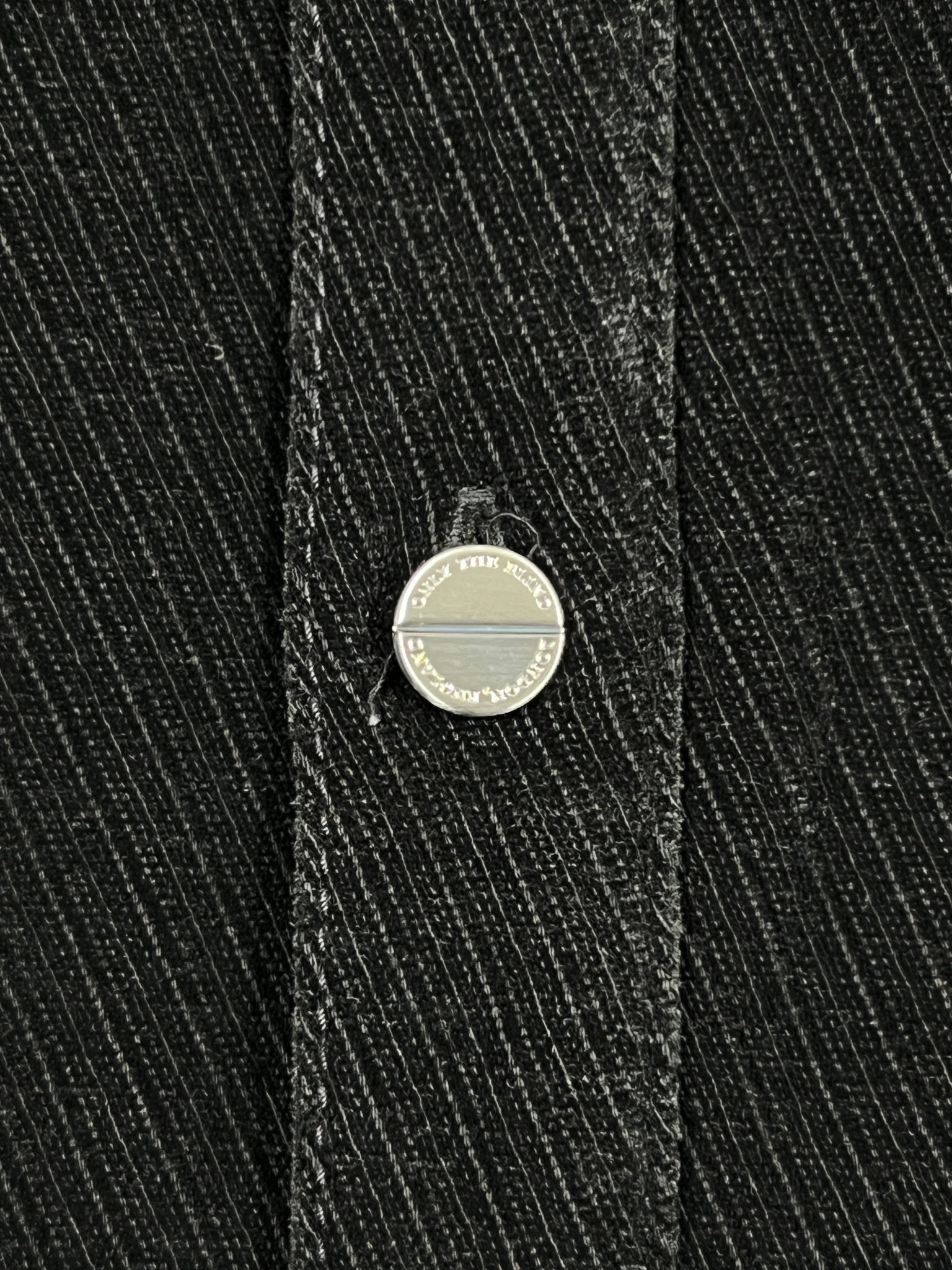Close-up of a metal button with engraved text on the edge, set on dark, diagonally striped fabric—part of the Probus Only The Blind OTB-SH1572 Noir Heritage Shirt Black.