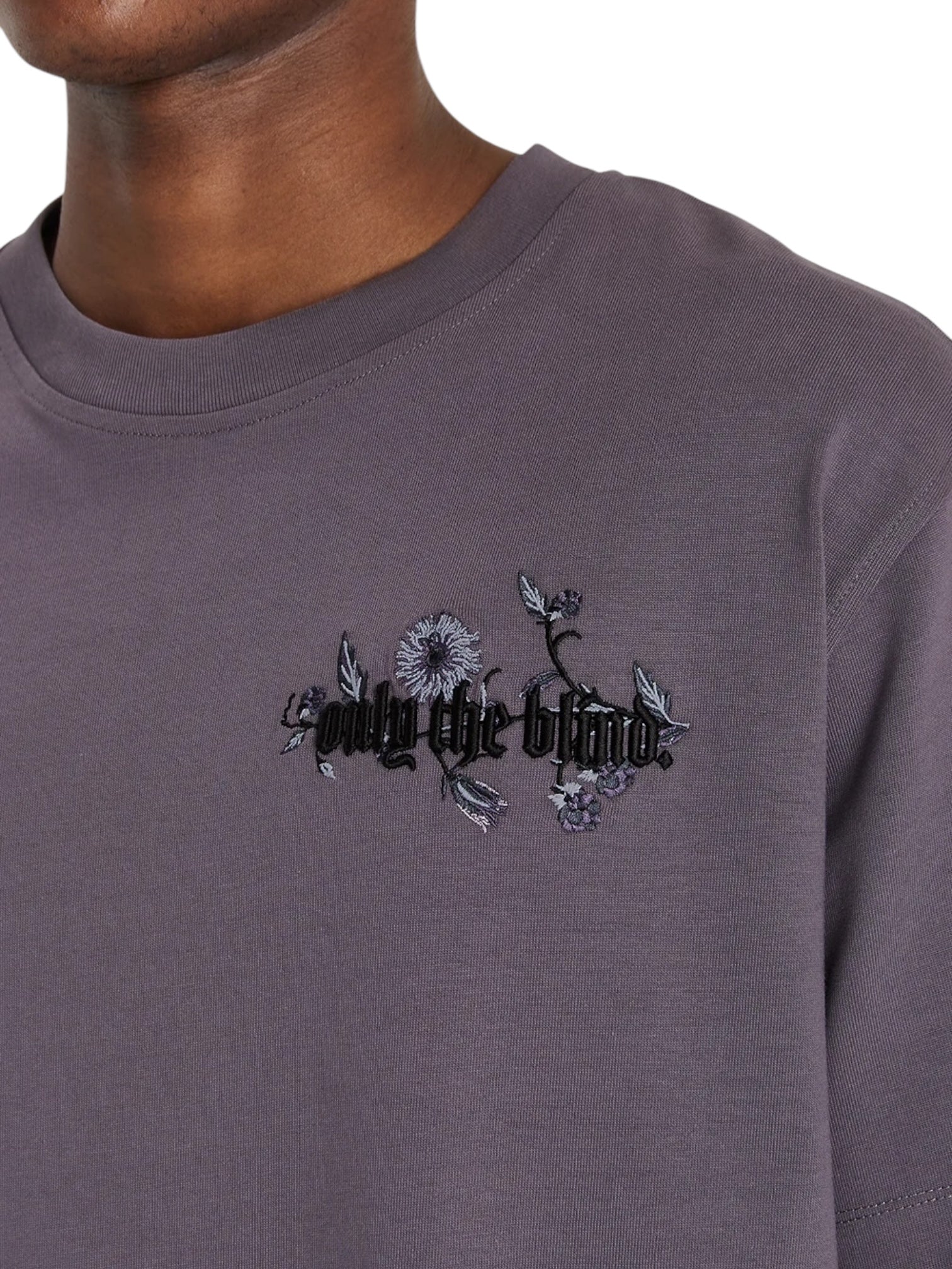 A person wearing an ONLY THE BLIND Wildflower T-shirt in purple, featuring blackberry floral embroidery and the text "only the blind" across the chest.