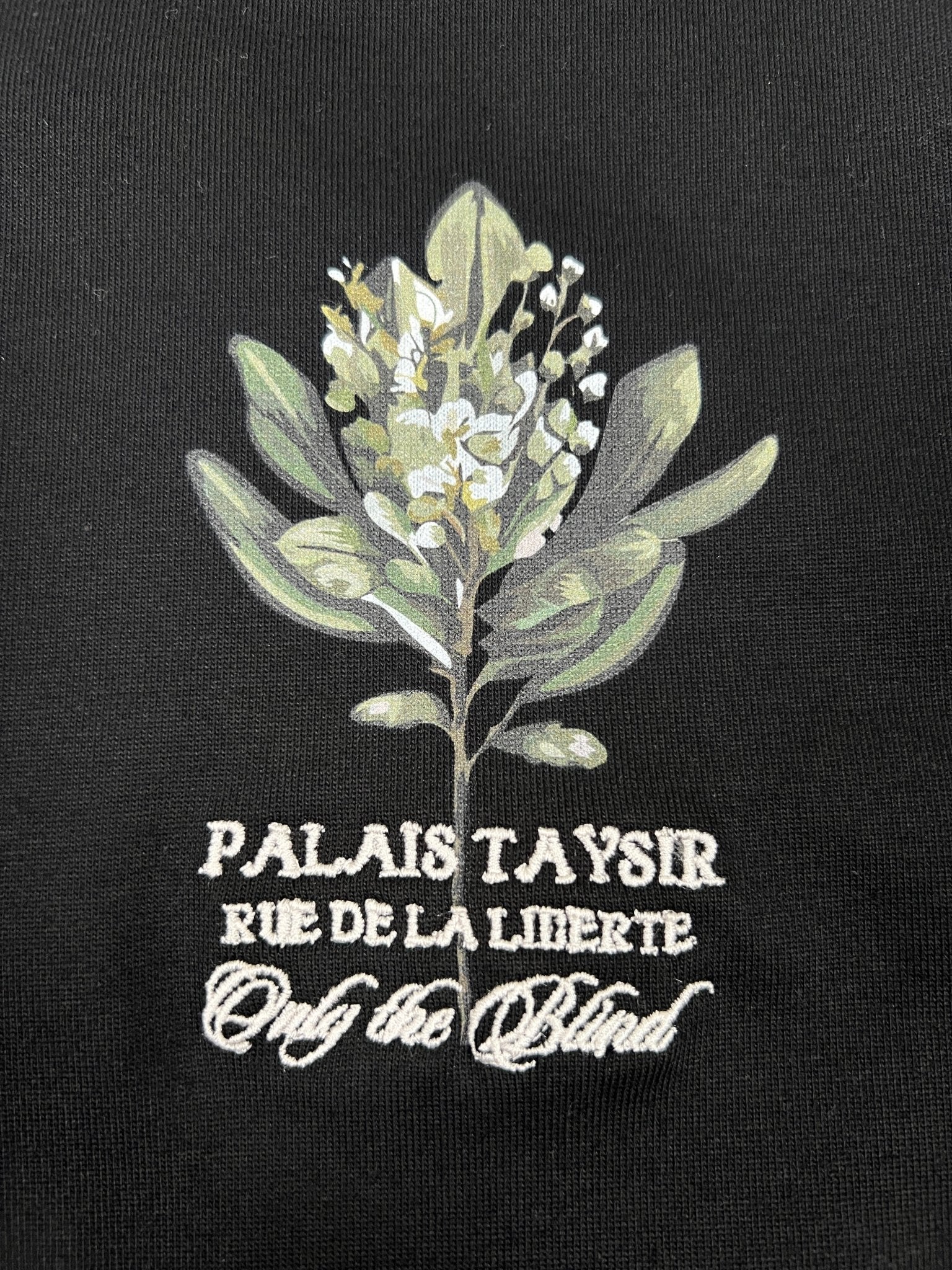 The ONLY THE BLIND OTB-T1472 Botanical T-shirt in black showcases an embroidered graphic of white flowers and leaves with "Palais Taysir, Rue de la Liberté, Only the Blind" text on soft organic cotton.