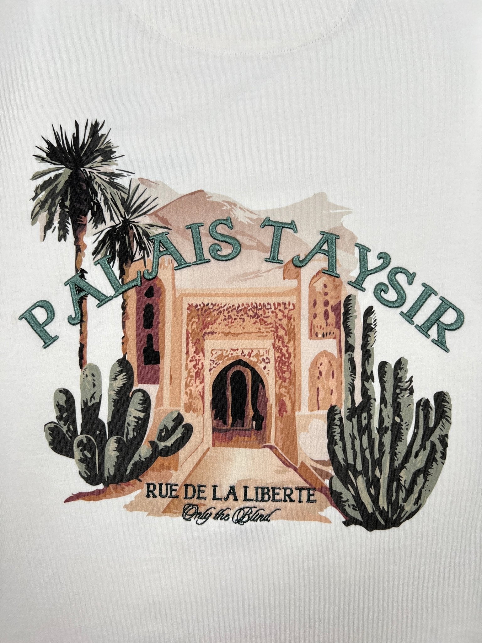 The ONLY THE BLIND OTB-T1486 Palais Taysir T-shirt Beige features an embroidered ornate arched building with palm trees and cacti, plus “PALAIS TAYSIR” and “RUE DE LA LIBERTE” captions on organic cotton.