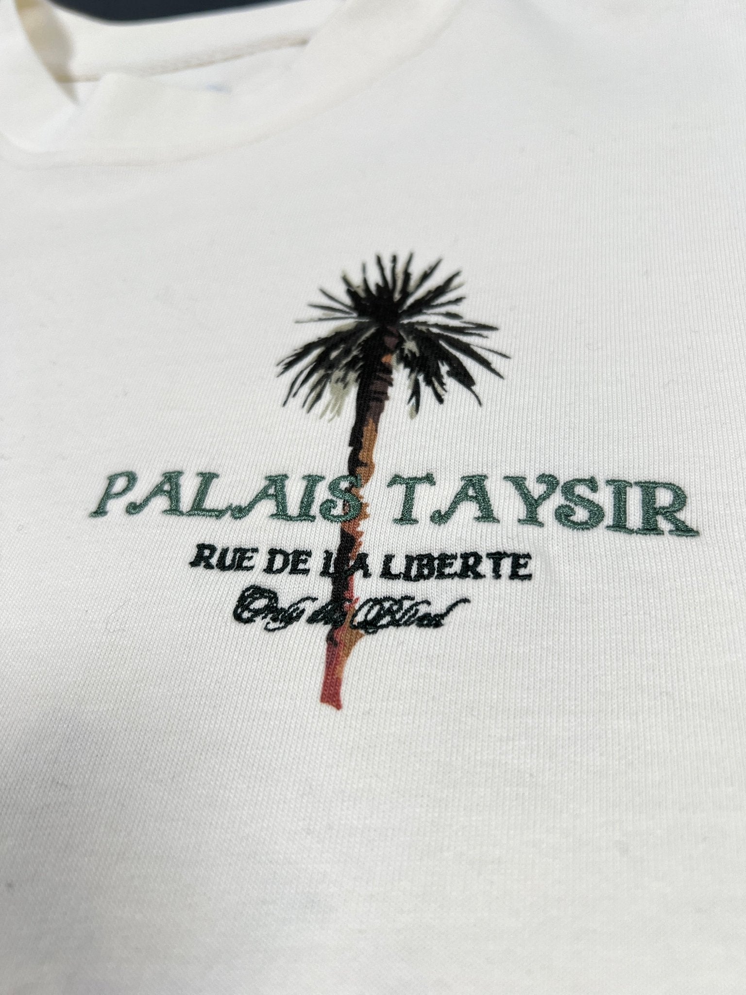 The ONLY THE BLIND OTB-T1486 Palais Taysir T-shirt in beige is made from organic cotton and features a white embroidered palm tree graphic with “PALAIS TAYSIR RUE DE LA LIBERTE” text in green and black.