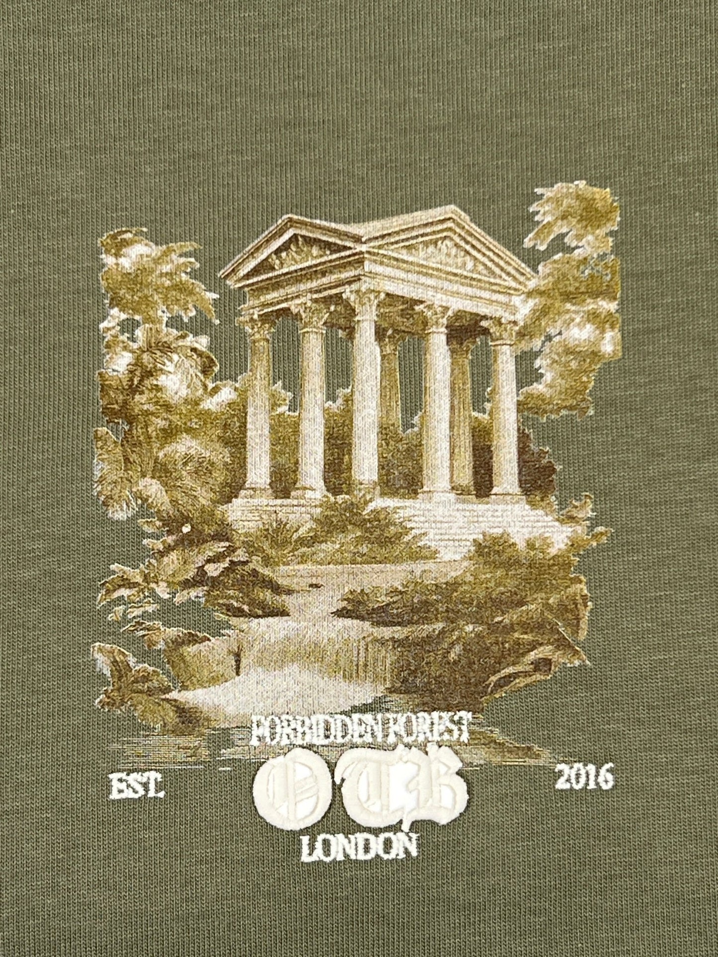 The ONLY THE BLIND OTB-T1589 Forbidden Forest T-shirt Green features an embroidered ancient Greek-style temple, trees, bushes, and white “FORBIDDEN FOREST OTHR EST 2016 LONDON” text on olive green 100% cotton.