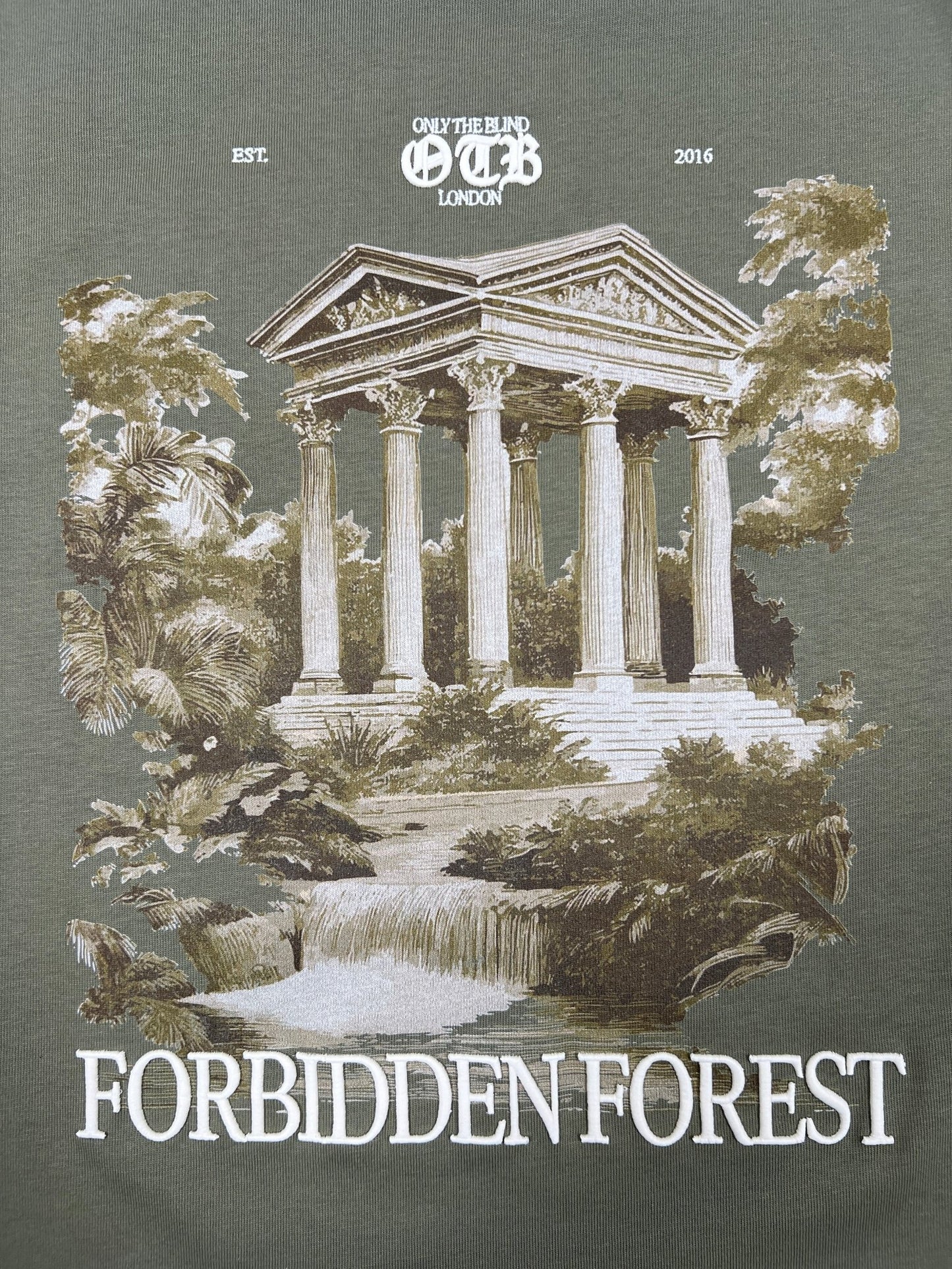 Illustration of a Greek-style temple, trees, and waterfall with “Only The Blind London” above and “Forbidden Forest T-shirt” below. The Only The Blind OTB-T1589 Forbidden Forest T-shirt Green is 100% cotton for style and comfort.