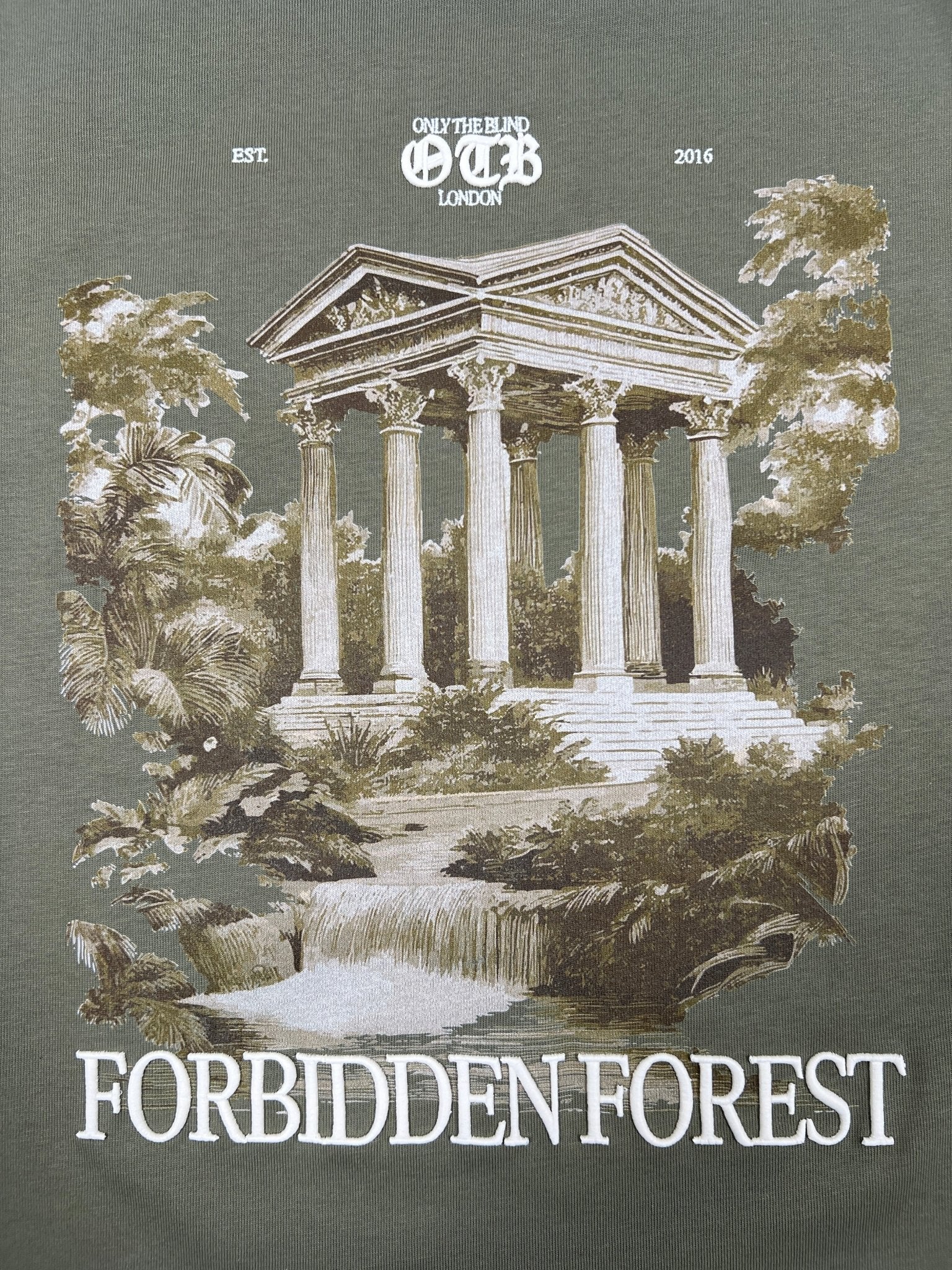 Illustration of a Greek-style temple, trees, and waterfall with “Only The Blind London” above and “Forbidden Forest T-shirt” below. The Only The Blind OTB-T1589 Forbidden Forest T-shirt Green is 100% cotton for style and comfort.