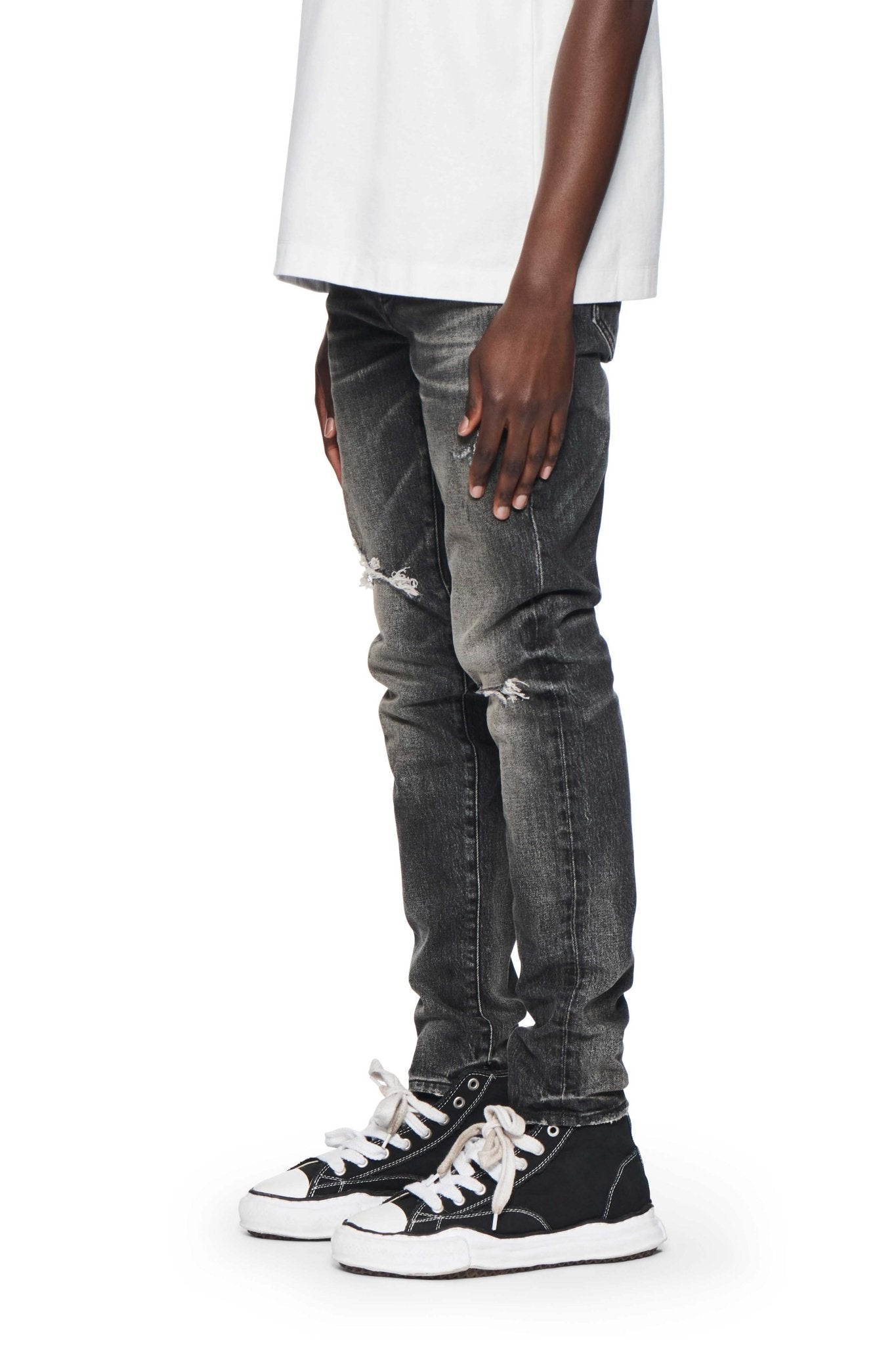 A person wearing a white shirt, faded dark grey ripped jeans (P001-SWKO SKINNY WEEK OLD WASH BLACK by PURPLE BRAND), and black high-top sneakers with white laces stands against a white background.