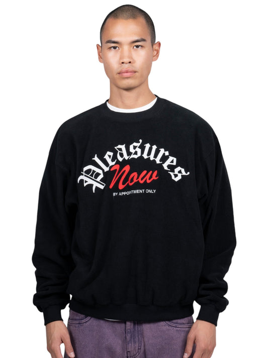 A person wearing the Pleasures Appointment Fleece Crew Black by PLEASURES, featuring "Pleasures Now" in bold white and red text, poses against a plain background.