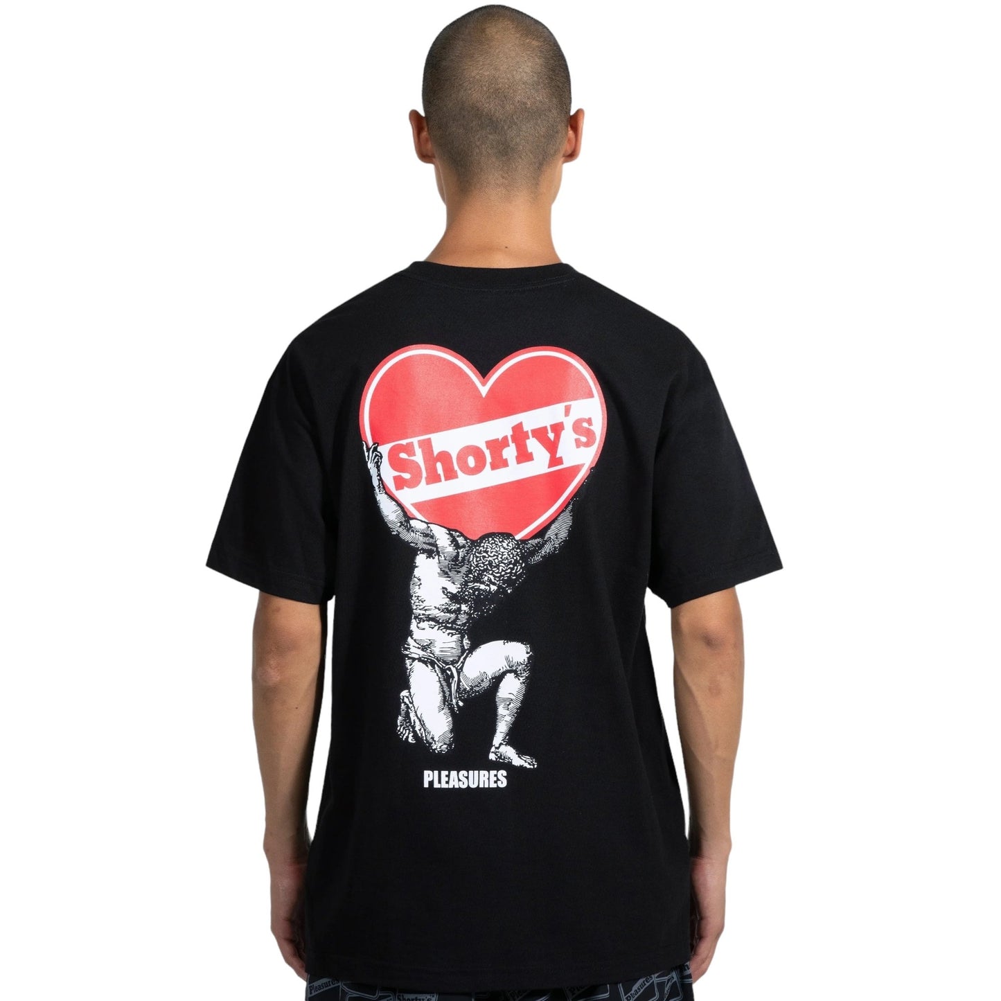 A person wearing the PLEASURES ATLAS T-SHIRT BLACK, made of 100% cotton, featuring 'Shorty's' printed inside a red heart on the back. Below the heart is an illustration of a figure holding it up with the word 'Pleasures' displayed underneath.