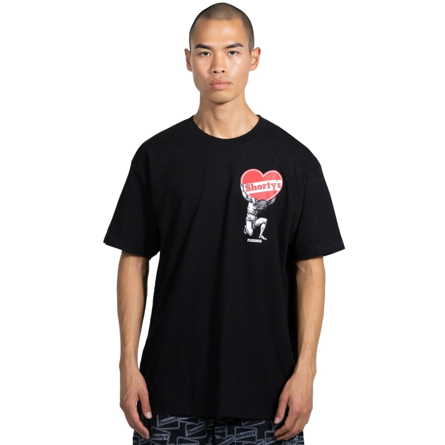 A man with a shaved head is wearing the PLEASURES Atlas T-Shirt in black, featuring a graphic of a figure holding a heart. This 100% cotton t-shirt from PLEASURES stands out against the plain white background.