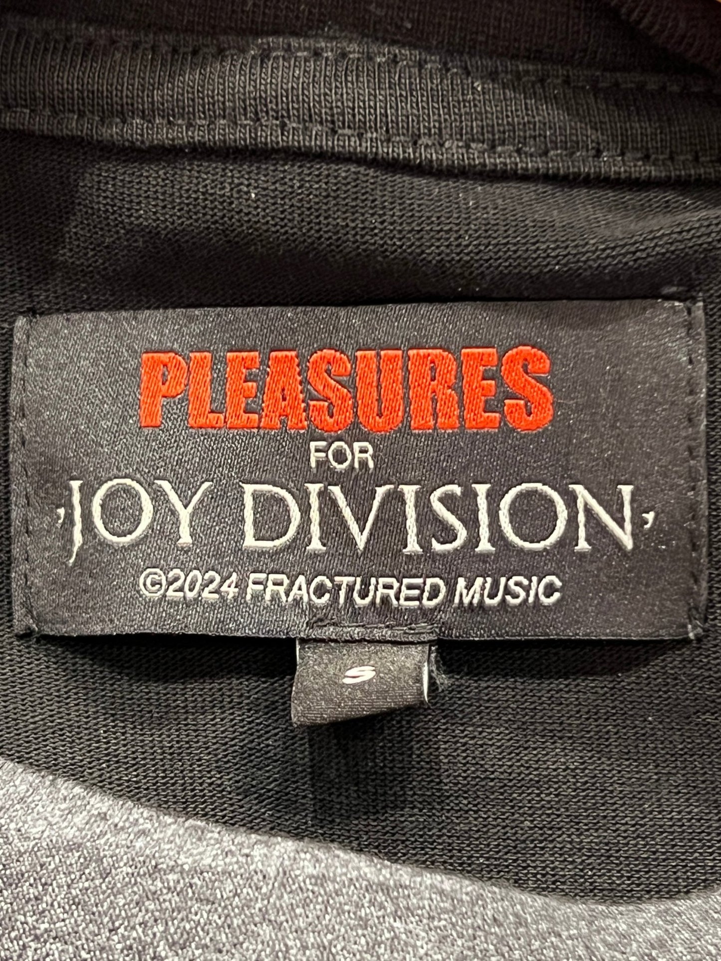Official collaboration Black cotton jersey PLEASURES ATROCITY TEE BLK clothing label with the text "pleasures for joy division ©2024 fractured music" in white and red font.