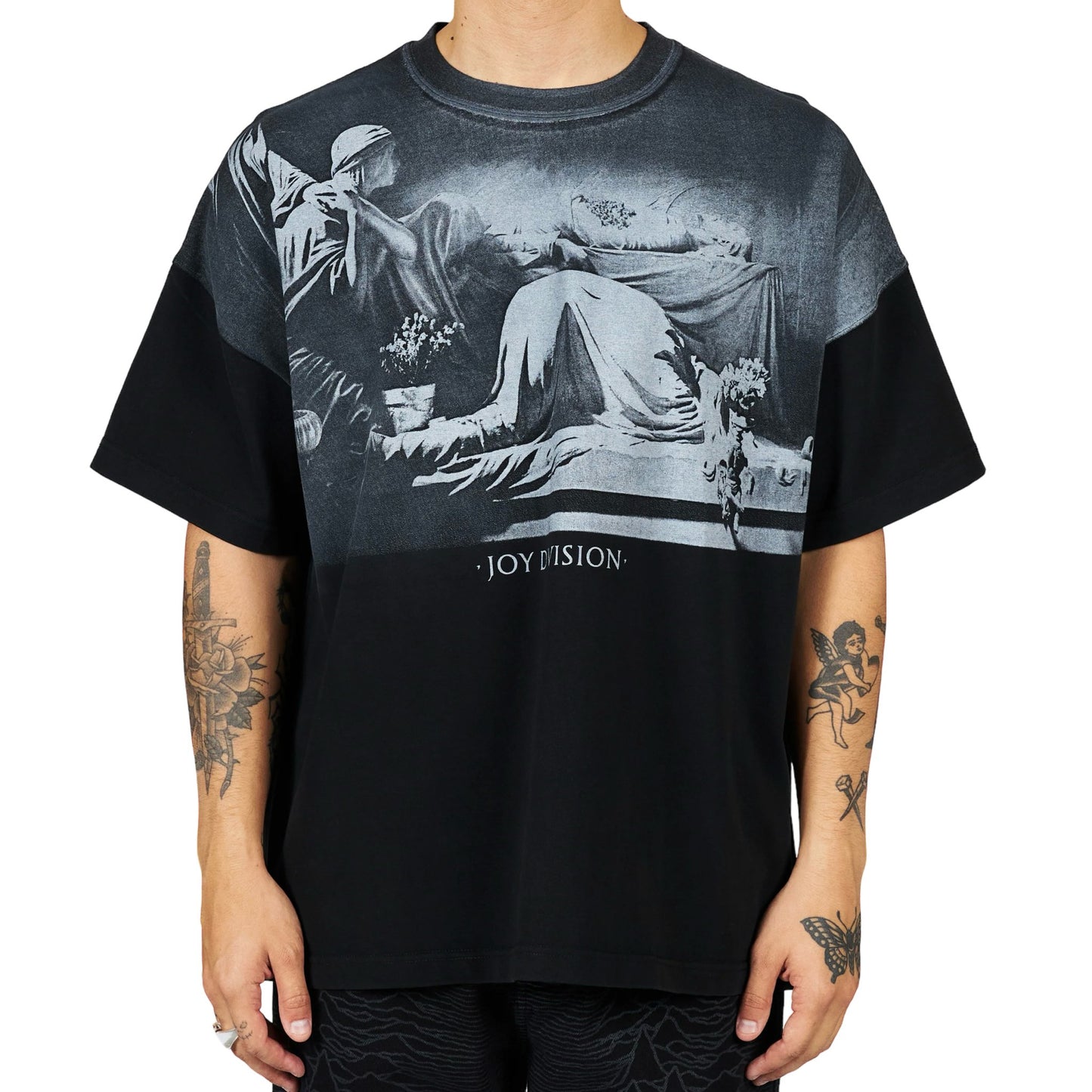 Man wearing a black t-shirt with an oversized plastisol print depicting a vintage scene and the text "Joy Division," an official collaboration with PLEASURES.