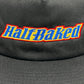 Close-up of the PLEASURES BAKED LOGO SNAPBACK BLACK, a hat made of Polyester & Nylon featuring "Half-Baked" embroidered in yellow, outlined in red and blue.