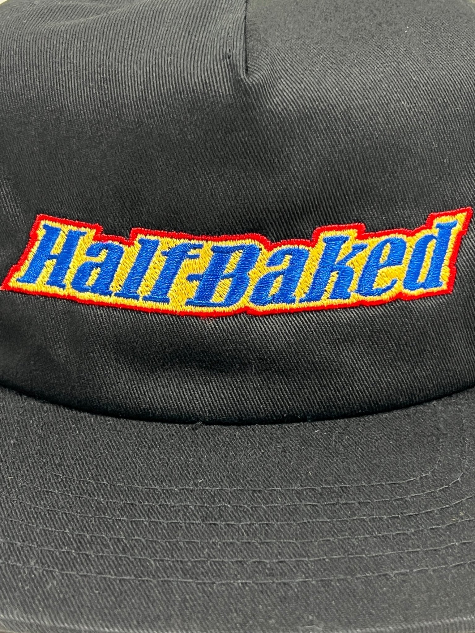 Close-up of the PLEASURES BAKED LOGO SNAPBACK BLACK, a hat made of Polyester & Nylon featuring "Half-Baked" embroidered in yellow, outlined in red and blue.