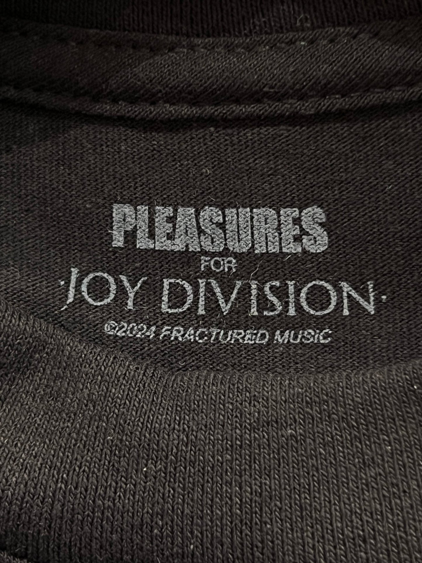 Black cotton fabric with PLEASURES BROKEN IN T-SHIRT BLK screen printed in white text, an official collaboration with PLEASURES.