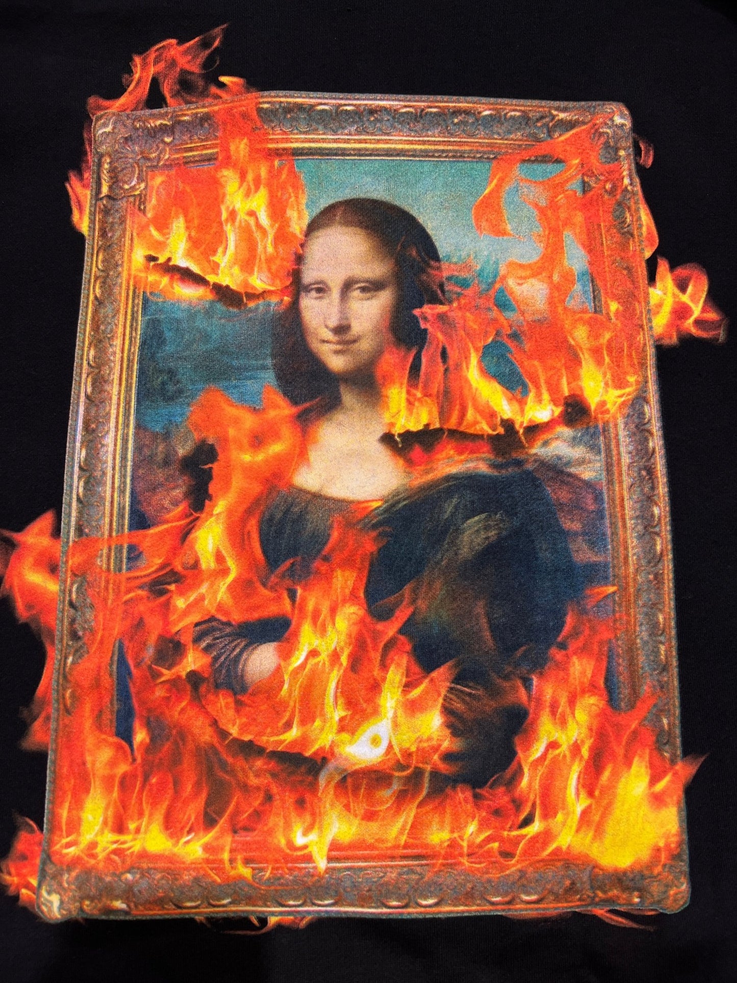 A captivating design of the Mona Lisa engulfed in vibrant flames is featured on the Pleasures Burn Again Hoodie Black by PLEASURES, creating an illusion of a tapestry of fire.