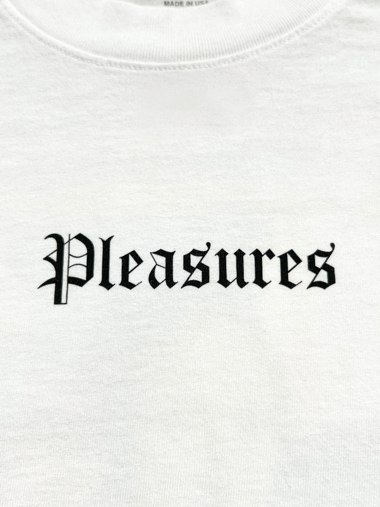 The Pleasures Burn Again T-shirt in white by PLEASURES features "Pleasures" in black Gothic-style print across the chest, crafted from garment dyed cotton for a soft, premium feel.