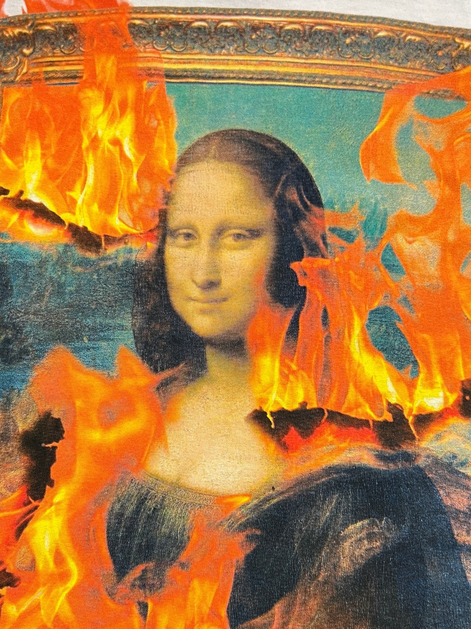 The Mona Lisa painting is partially obscured by vivid flames, giving the artwork a bold twist—just like the PLEASURES Burn Again T-shirt White, which stands out with its striking, boxy fit design.