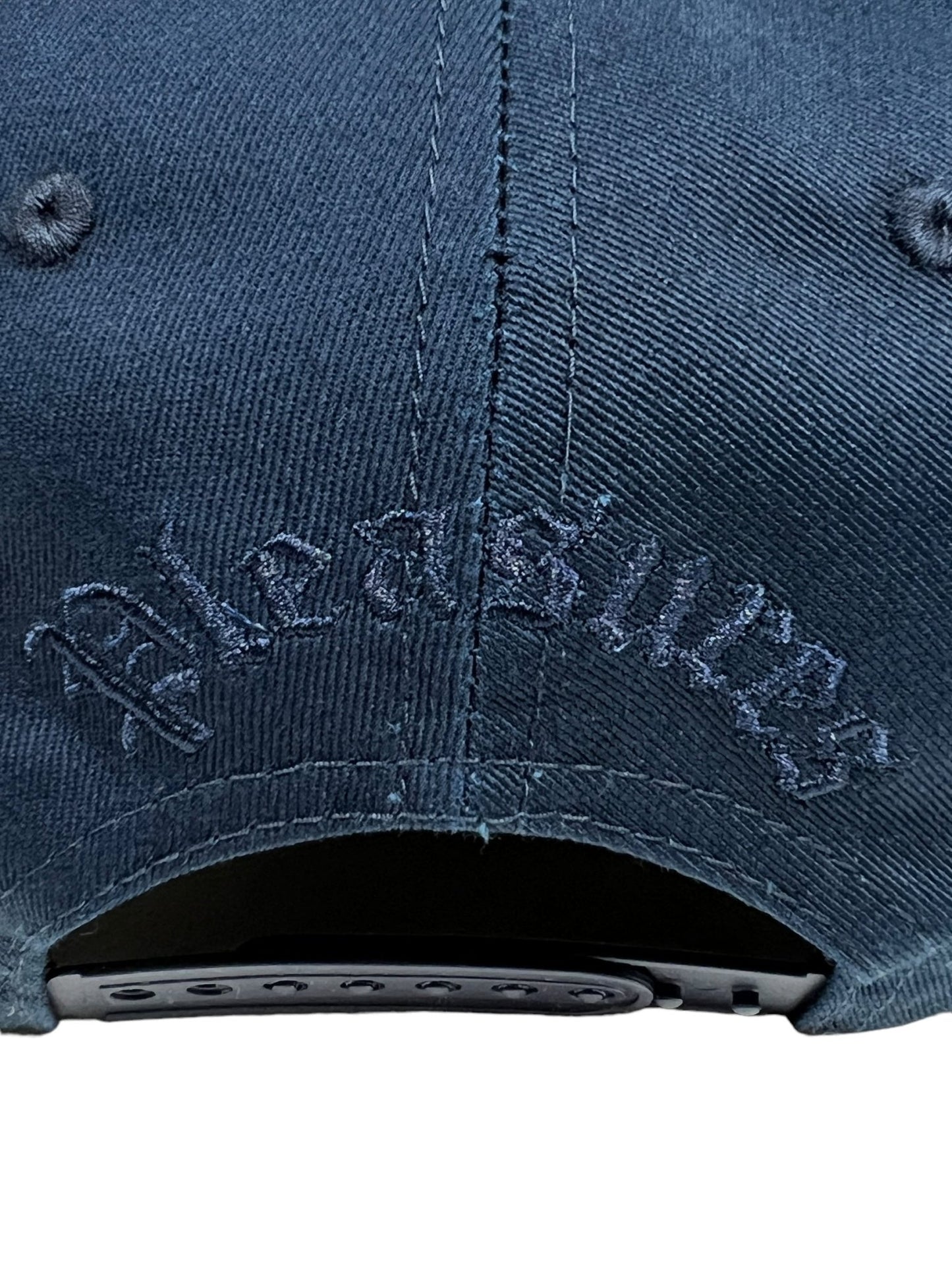 Close-up of the back of the PLEASURES CHROME SNAPBACK NAVY hat by PLEASURES, showcasing a plastic adjustable strap and the word "PLEASURES" embroidered in matching navy blue thread.