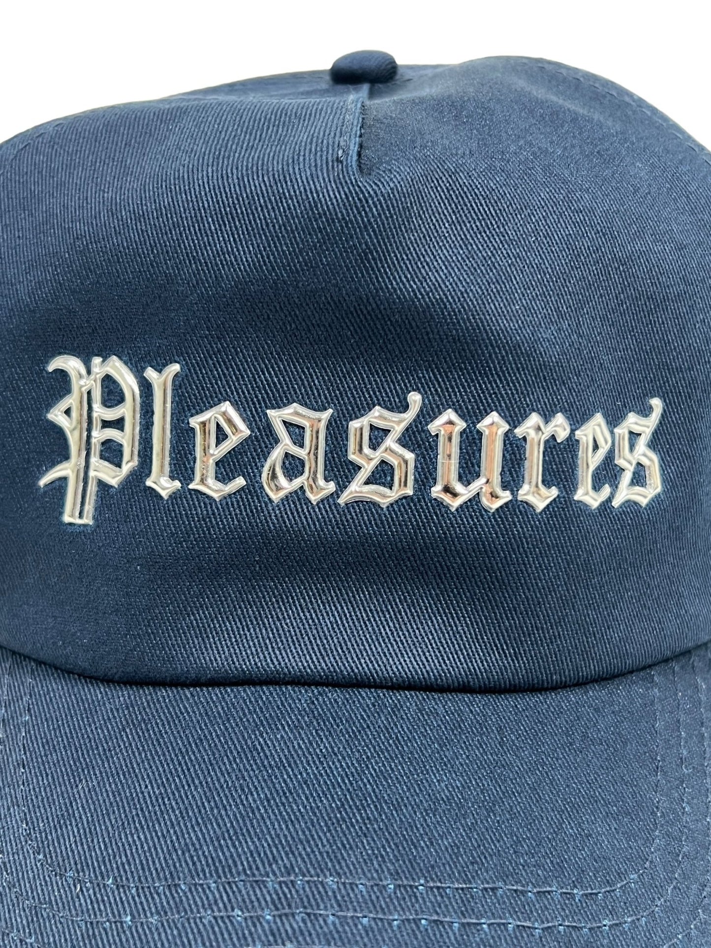 Close-up of the PLEASURES CHROME SNAPBACK NAVY, a navy blue hat made from Polyester & Nylon, featuring the word "Pleasures" written in Gothic-style white letters on the front.