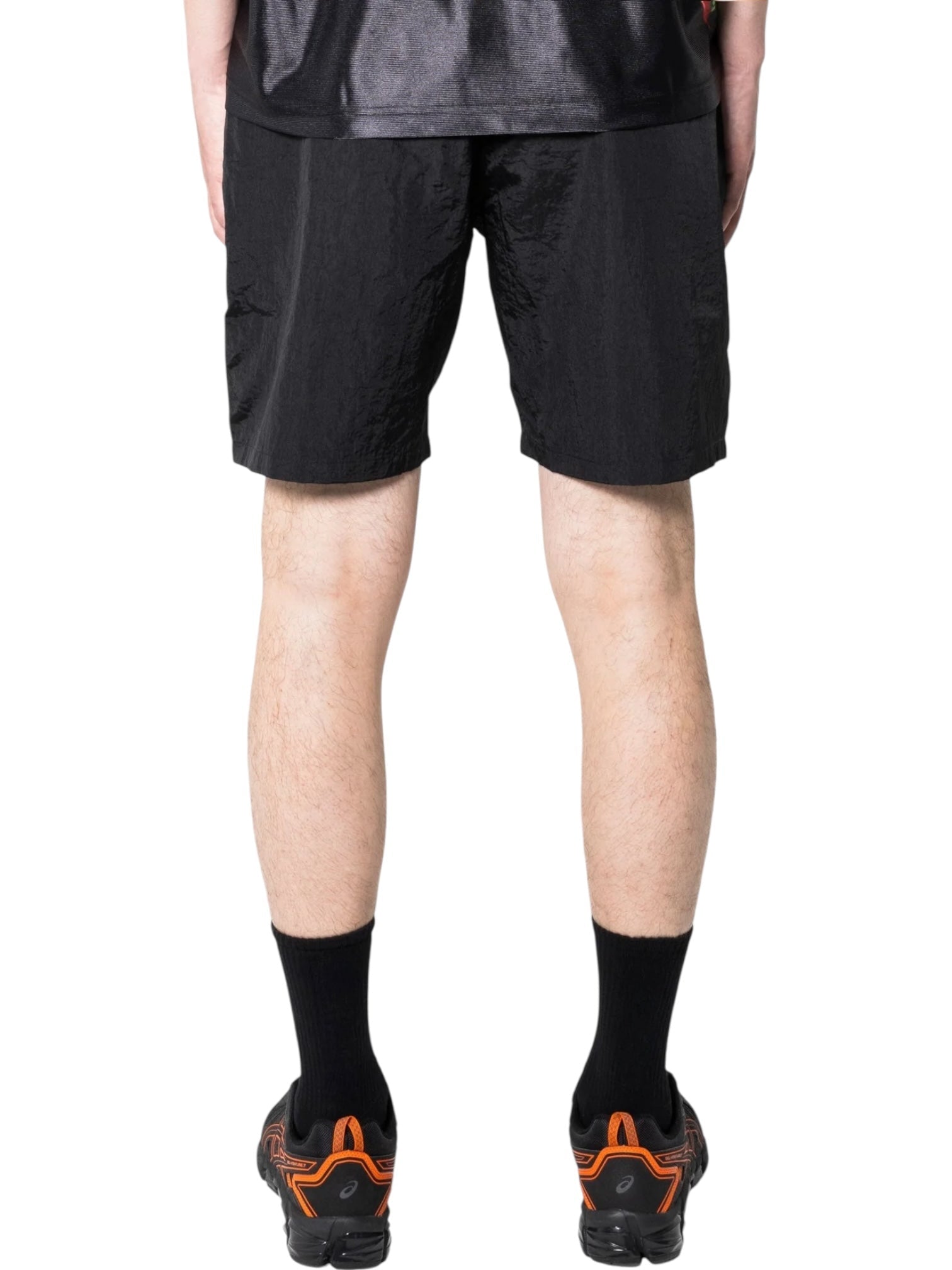 A person shown from the back wears a black shirt, PLEASURES Pleasures Crush Hiking Shorts Black with an elasticized waistband, black crew socks, and black athletic shoes with orange accents.