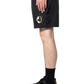 A person in PLEASURES Crush Hiking Shorts Black with an elastic waistband and circular patch, paired with black socks and black sneakers with orange details, stands in profile against a white background.