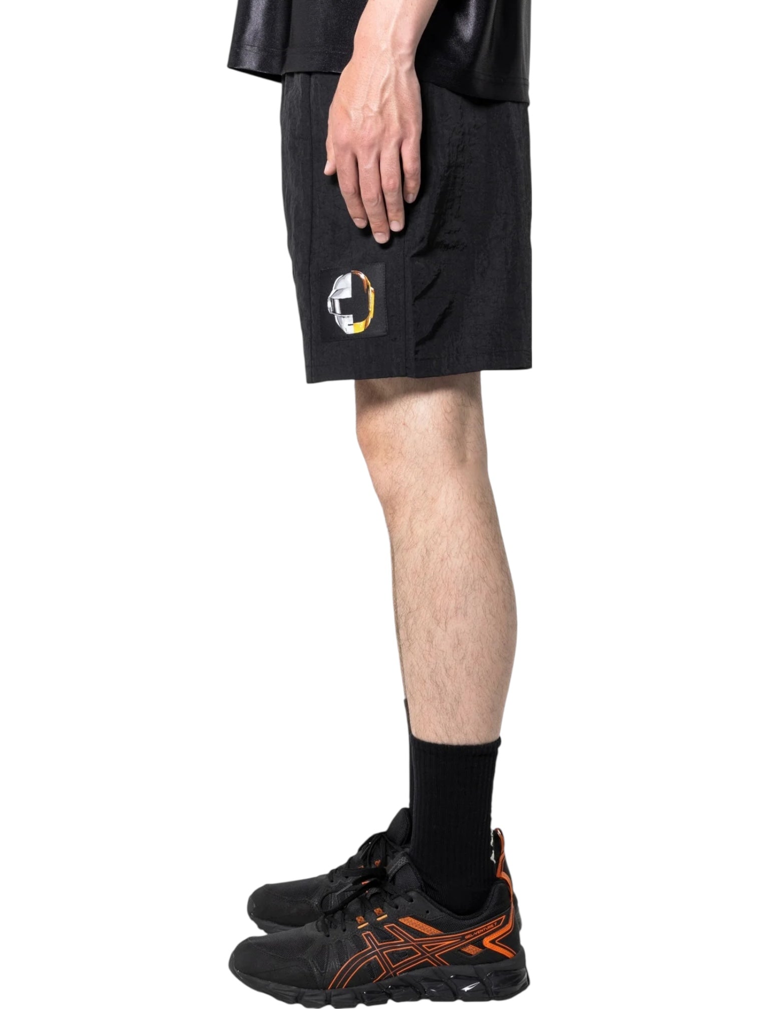 A person in PLEASURES Crush Hiking Shorts Black with an elastic waistband and circular patch, paired with black socks and black sneakers with orange details, stands in profile against a white background.