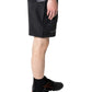 A person stands in profile wearing PLEASURES Crush Hiking Shorts Black with an integrated belt, a black shirt with a colorful design, black socks, and black sneakers featuring orange accents.